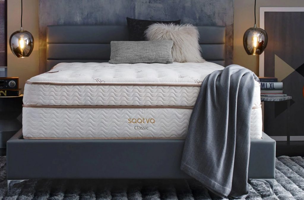 Front view at eye level of The Saatva Classic Mattress, showing the organic fabric used on the pillow top, the gold contrasting embroidery, and a draped grey throw across a corner of the mattress
