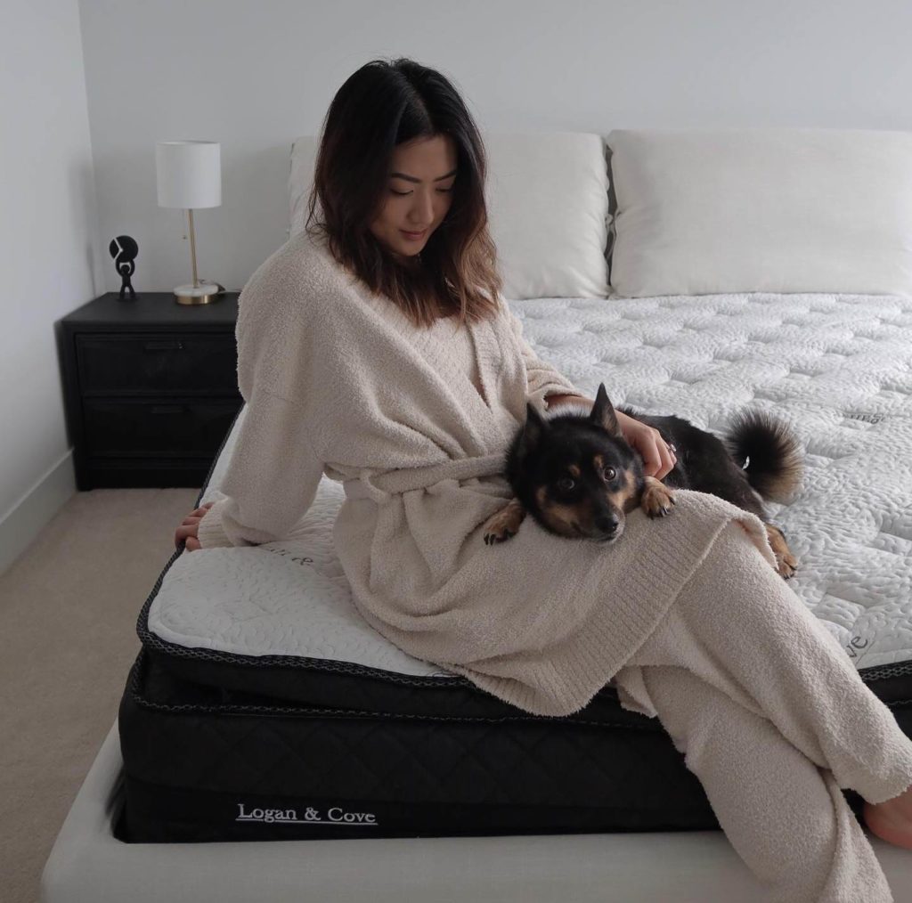 A best mattress Canada option 
depicts woman in bathrobe near edge of Logan & Cove Mattress. She is petting a small dog, image is overhead shot offering a comforting view. Pillows and nearby nightstand are also in view, darker version