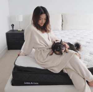 Relaxing woman in bathrobe near edge of Logan & Cove Mattress. She is petting a small dog, image is overhead shot offering a comforting view. Pillows and nearby nightstand are also in view