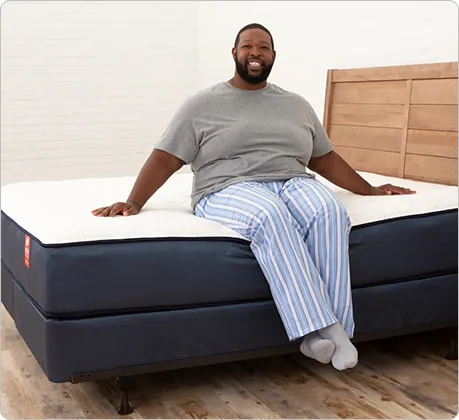 Image shows a large man sitting on the edge of a mattress. The photo demonstrates the importance of edge support and ability to transfer easily from the edge of the mattress . This mattress is called BigFig and is designed with firmer edge support for people that have bigger bodies.