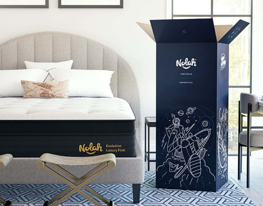 photo showing a nolah evolution mattress placed on a bed frame with pillows, and next to it is the opened shipping box which is about 26x26x48 inches tall. this illustrates the manageable sized box that is provided during shipment.