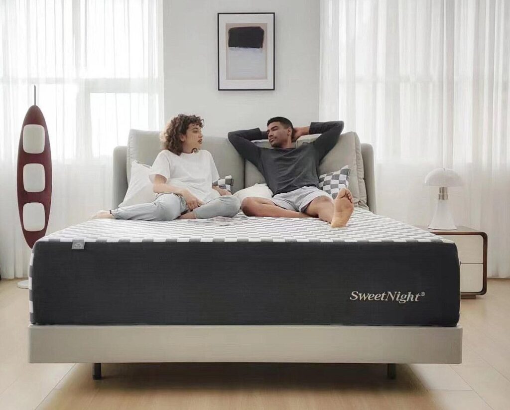 Photo shows a young couple reclined on the Sweetnight Prime Mattress. The room is soft and white with curtains agains the windows. The photo shows the couple from the foot of the mattress.